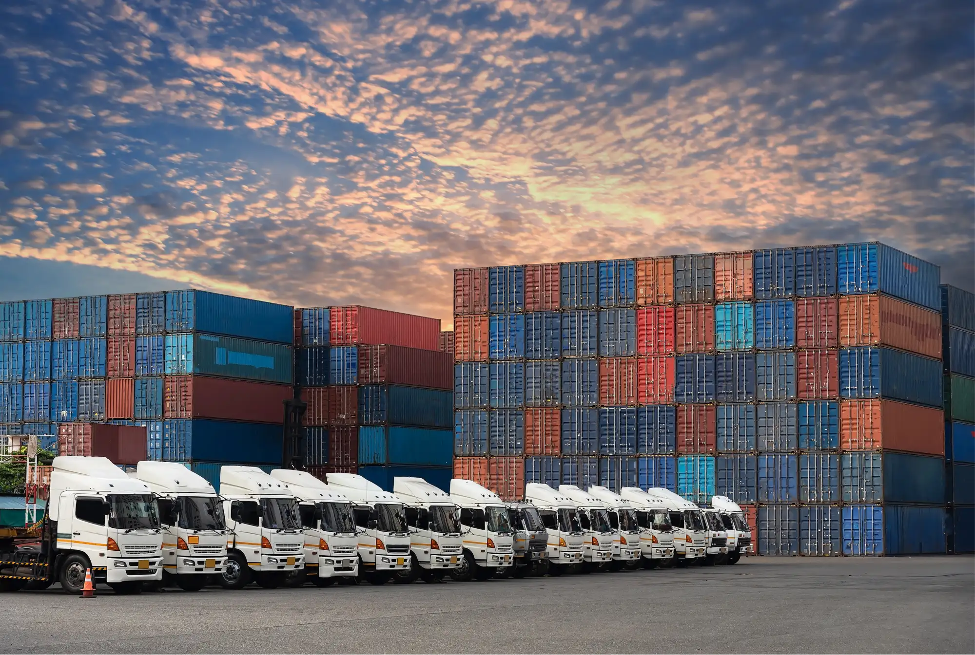 Fleet of freight trucks parked in front of stacked shipping containers at dusk